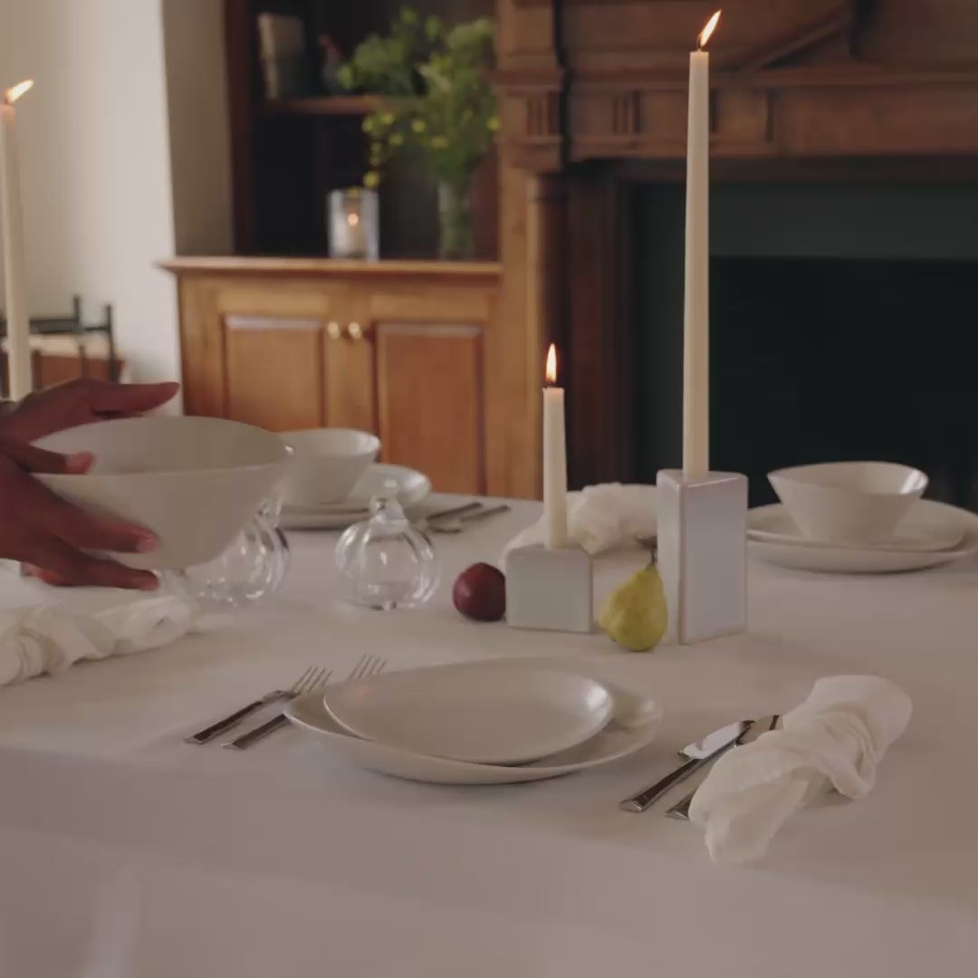 Hands setting a table with white dinnerware on a white linen runner with candles flickering in white pottery candle holders