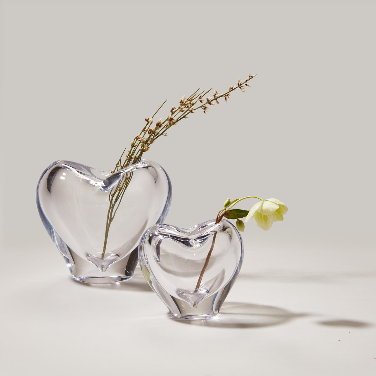 Two clear glass heart-shaped vases holding dried botanicals and fresh greenery.