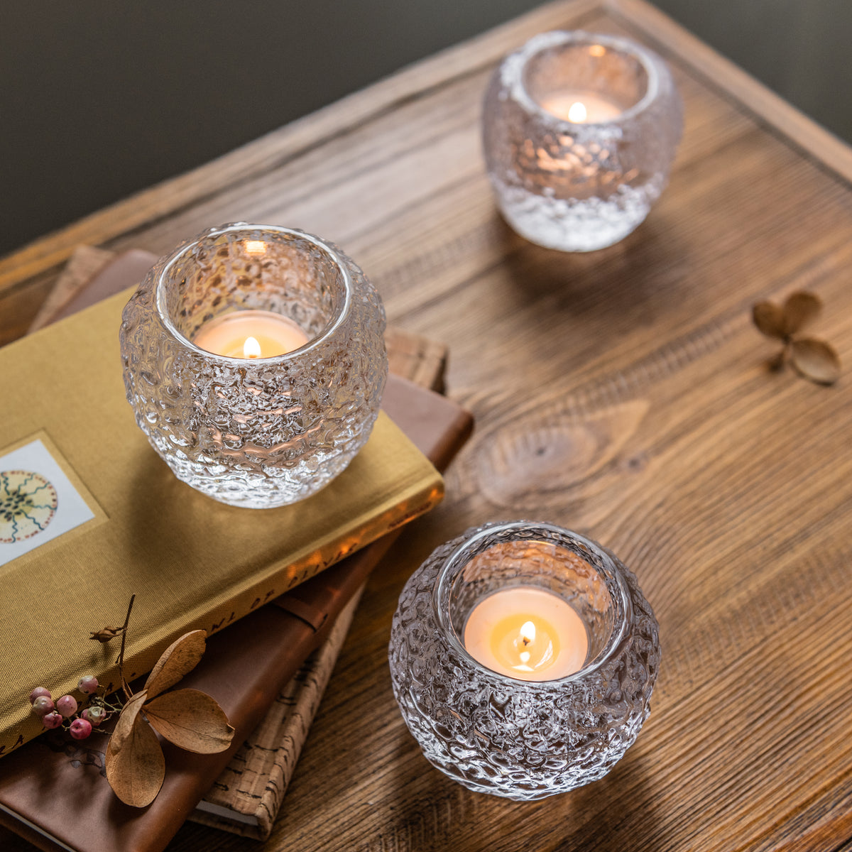 Three textured glass tealight holders with lit candles arranged on a wooden surface with decorative items.