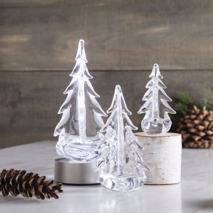 Three clear glass evergreen tree figurines with metal and birch wood bases, arranged with pinecones on white surface.