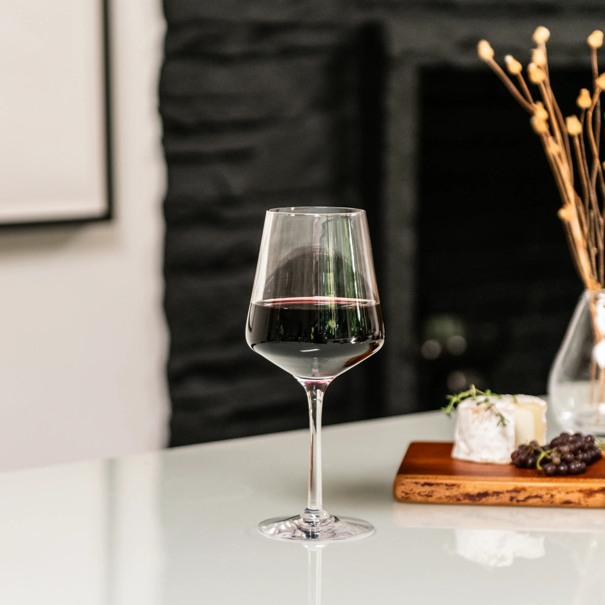 Red wine in a stemmed glass on white table with cheese, grapes, and decorative elements in background.