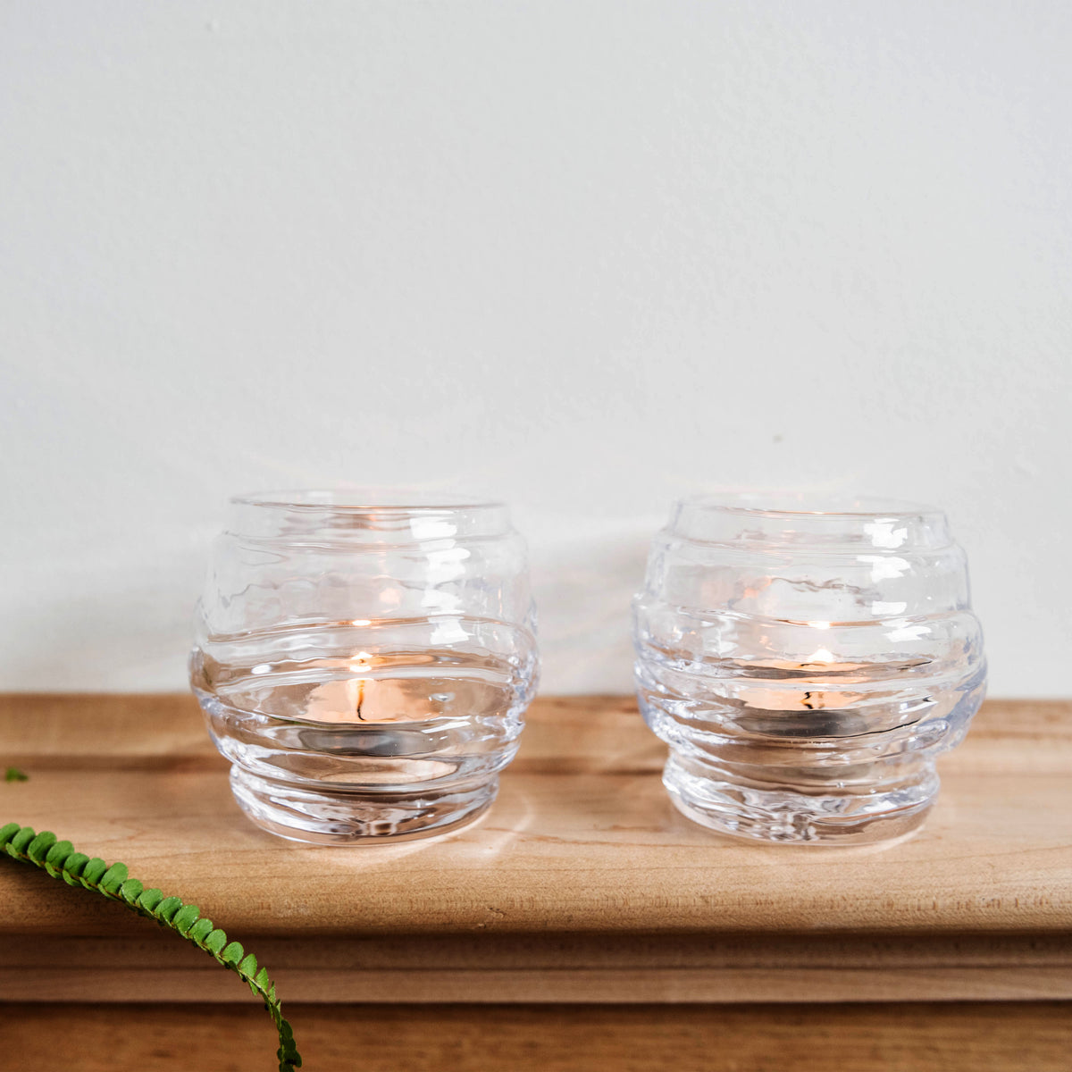 Two clear glass tealight holders with horizontal ribbed pattern, one containing a lit candle, displayed on wooden tray.