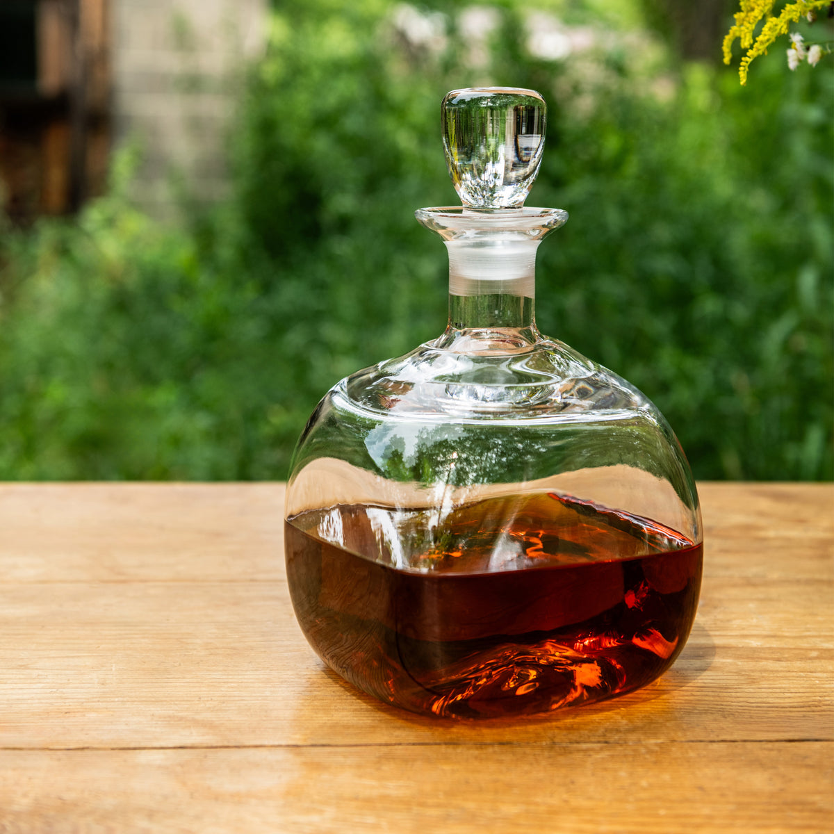 Clear glass decanter with amber liquid and matching stopper, displayed on wooden surface outdoors.