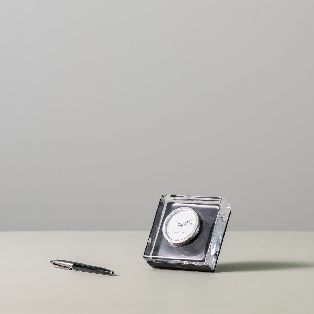 Square clear acrylic clock with white face and round dial in center, displayed next to pen for scale.